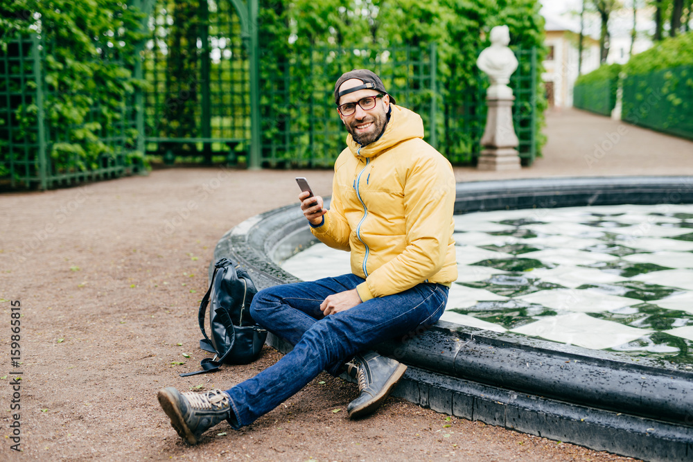 Smiling happy man with bristle wearing casual clothes and big eyewear sitting crossed legs near fountain in park holding cell phone messaging with his girlfriend having delightful expression