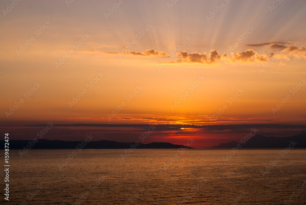 Horizontal image of golden sunset with sun low above the sea covered by clouds and sunrays coming through clouds above
