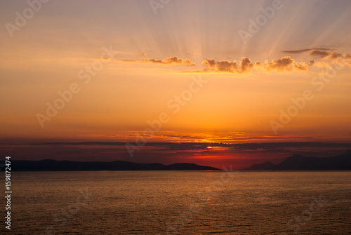 Horizontal image of golden sunset with sun low above the sea covered by clouds and sunrays coming through clouds above  