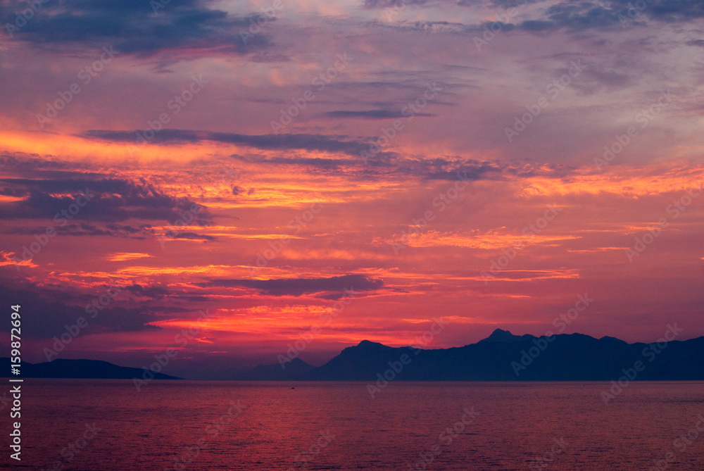 Horizontal image of deep coloured sunset with sea in red, golden and purple colours, black mountains on the horizon,