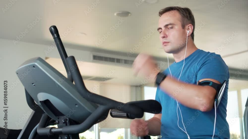 Fit young man in gym doing exercises on velosimulator. Listening music on smartphone with ear-phones Fit young man in gym doing exercises on velosimulator. Listening music on smartphone with ear-phones