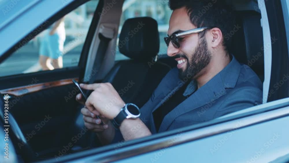 Handsome bearded man in stylish sunglasses sits in the business class car holding the phone, gets the message, smiles happily and respond to it. Successful lifestyle, luxurious look, corporate style. Handsome bearded man in stylish sunglasses sits in the business class car holding the phone, gets the message, smiles happily and respond to it. Successful lifestyle, luxurious look, corporate style.