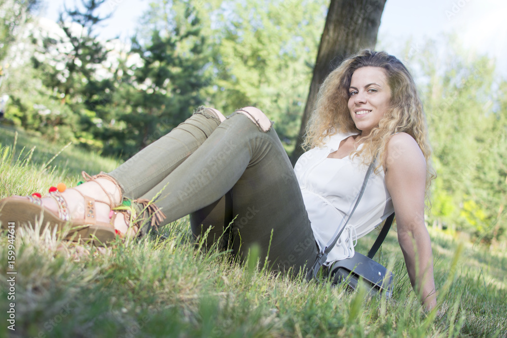 Fototapeta premium Gorgeous young curly-haired woman relaxing at the city park. Woman in nature, trees in the background.