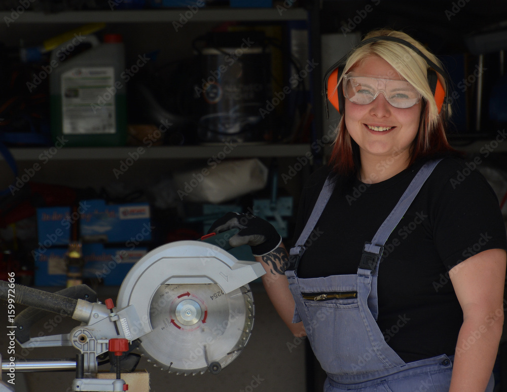 Fototapeta premium Junge lächelnde Frau im Overall mit Gehörschutz und Schutzbrille an Kreissäge