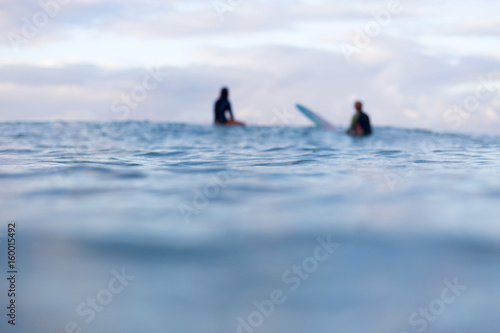 Two out of focus surfers sit in the distance on their surfboards as they wait for a wave to appear on the horizon.