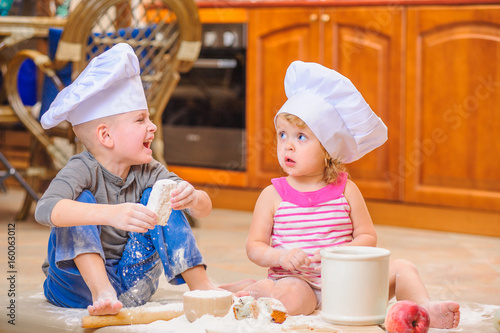 Two siblings - boy and girl - in chef's hats sitting on the kitchen floor soiled with flour, playing with food, making mess and having fun