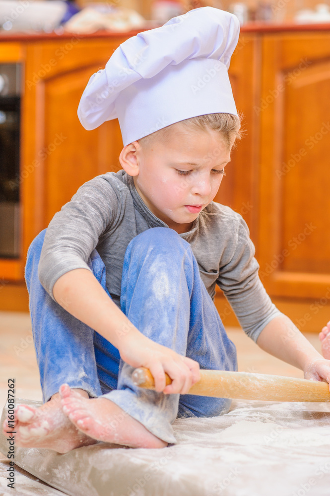 Two siblings - boy and girl - in chef's hats sitting on the kitchen floor soiled with flour, playing with food, making mess and having fun