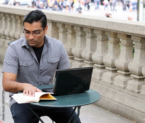 handsome man reading book while surfing the web on laptop in an outdoor urban park photo