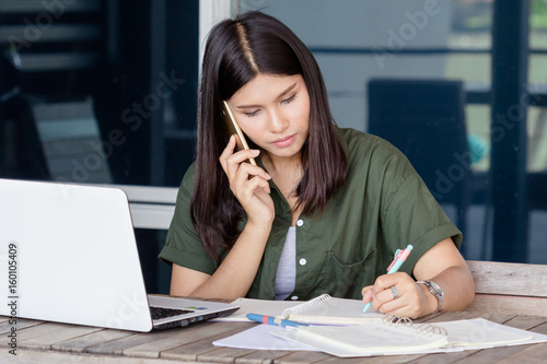 beautiful asian woman writing and talk smartphone on the wooden table with notebook pen papersheet smartphone at workplace with sun light working concept photo