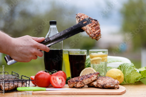 Man's hand holds a Chicken breast on the grill tongs on a wooden Board. , Food photography chicken chili steak on the background with vegetables and black soda and lemonade photo