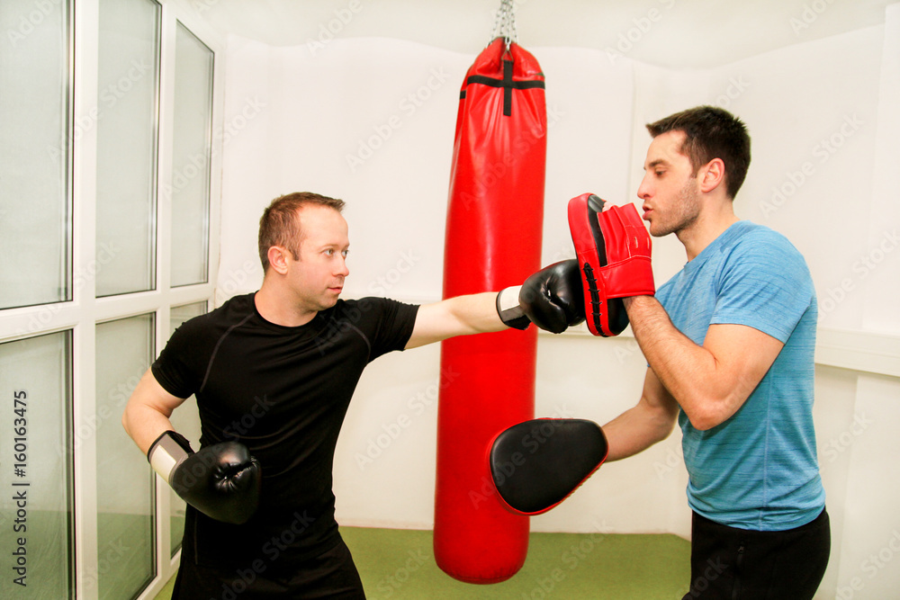The male boxer training at gym. Athletic man punching her male partner and instructor with red boxing gloves at gym club. Attractive sports man is practicing with a handsome trainer in sport center. The male boxer training at gym. Athletic man punching her male partner and instructor with red boxing gloves at gym club. Attractive sports man is practicing with a handsome trainer in sport center.