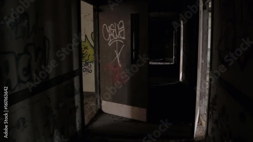 FPV, CLOSE UP: Walking along dark narrow hallway in abandoned decaying psychiatric hospital. Moving past the weathered wooden doors and broken crumbling walls in sinister sanatorium looking for escape photo
