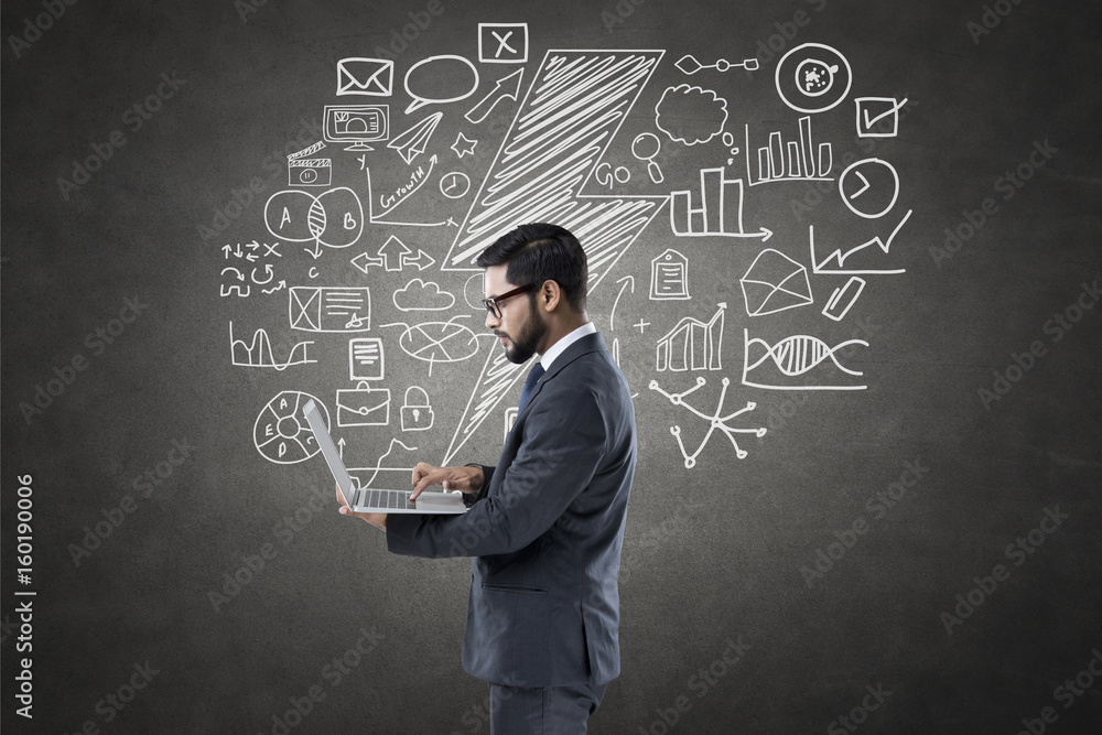 Businessman using laptop in front of blackboard with icons Businessman using laptop in front of blackboard with icons