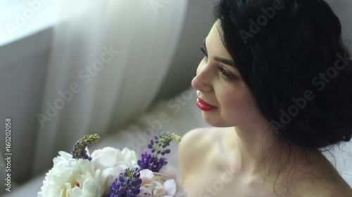 The morning of a gentle attractive brunette. Beautiful bride is smiling and looking out the window and a bouquet while sitting on the bed.