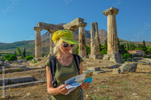 Greece tourist woman reads travel guide outdoors. Happy female holding informative guide at Doric Temple of Apollo in Ancient Corinth, Peloponnese. Acrocorinth on background. Tourism people concept.
