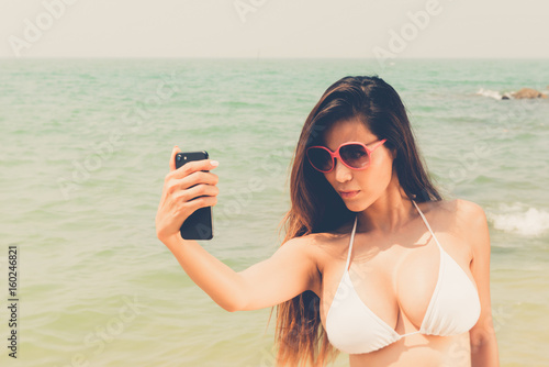 Beautiful attractive large breast asian bikini woman posing sexy portrait on beach smart phone selfie