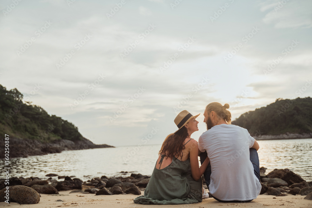 Young loving guy with girl sitting on beach beach at sunset and looking at each other. Love story. Concept of relaxation and romance.