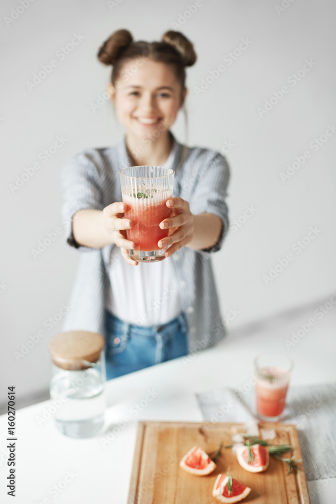 Happy girl with buns smiling stretching grapefruit detox smoothie to camera over white wall. Healthy diet food. Focus on glass. Happy girl with buns smiling stretching grapefruit detox smoothie to camera over white wall. Healthy diet food. Focus on glass.
