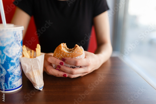 A girl holds a hamburger in a cafe on the background of the drink on the wooden table in fast food dining