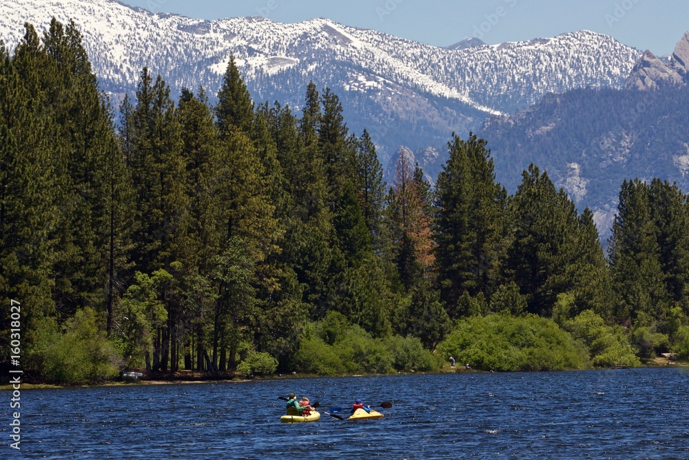 Paddlers on a Mountain Lake - paddlers paddle two kayaks on a blue mountain lake with green forest and snow covered mountains in the background