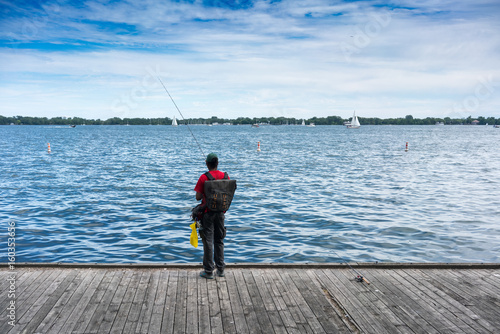 Person standing on pier fishing holding fishing rod wearing backpack rear view Ontario Canada