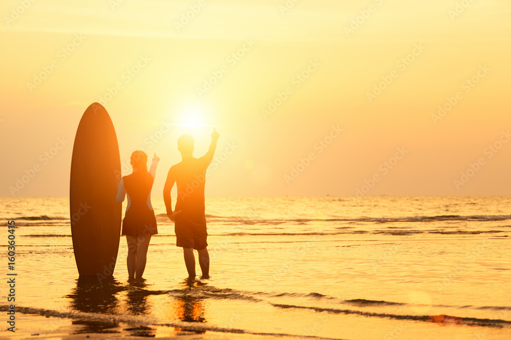 Silhouette of a man, a woman relaxing near tanding with surfboards on the beach at sunset time.