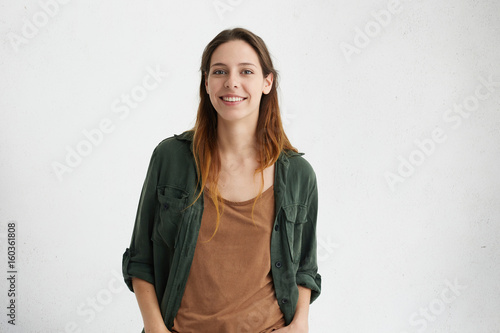 Pleased pretty woman with dyed hair, dark eyes and healthy skin dressed in brown T-shirt, green jacket holding hands in pockets smiling while posing against white concrete wall. People and lifestyle