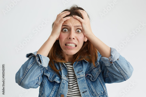 Portrait of shocked terrified woman with dark eyes popped out clenching her teeth holding hands on head looking in despair into camera isolated over white background. Woman having frustration photo