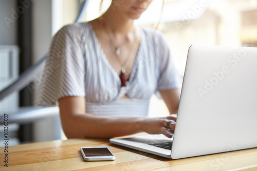 Free Download Cropped view of businesswoman sitting at wooden desk indoors surrounded with modern gadgets. Clever woman working online via laptop computer typing report. Woman's hands keyboarding on computer - GFXLoad