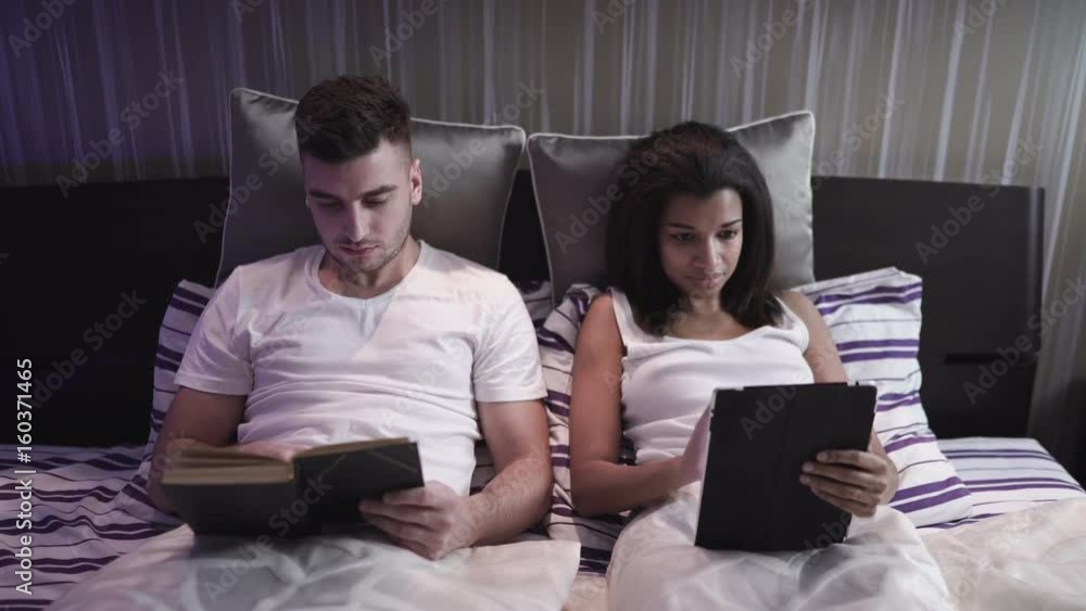 Young couple in modern bedroom, man reads a book and the woman looks in her pc tablet Young couple in modern bedroom, man reads a book and the woman looks in her pc tablet