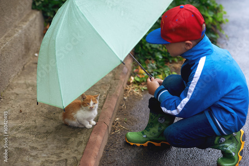 the child taking care of a kitten on the street , an umbrella sheltering him from the rain .