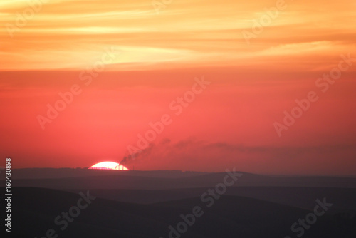 Large sun setting over the horizon in the hills covered with mist with smoking chimneys