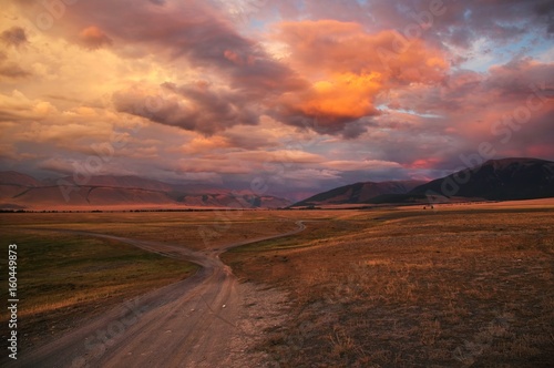 Road path on a desert wild mountain plateau at the background of the hills under a dramatic sunset colorful sky with illuminated red pink purple clouds Kurai Altai Siberia Russia