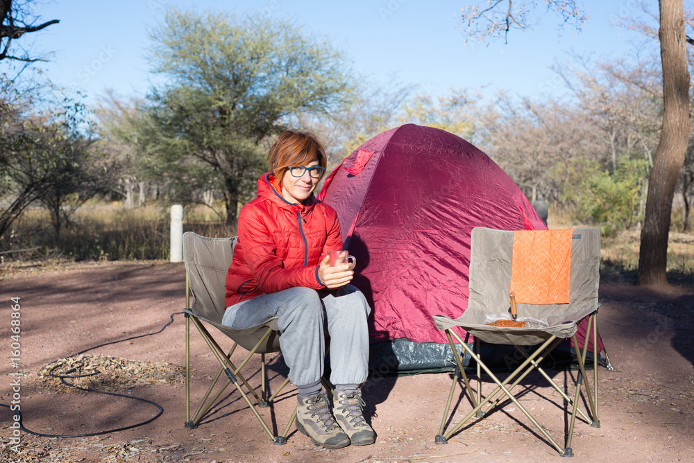 Woman drinking hot coffee mug while relaxing in camping site. Tent, chairs and camping gears. Outdoor activities in summer. Adventure traveling in South African National Park. Woman drinking hot coffee mug while relaxing in camping site. Tent, chairs and camping gears. Outdoor activities in summer. Adventure traveling in South African National Park.
