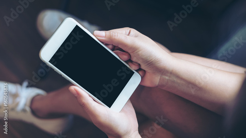 Fototapeta Naklejka Na Ścianę i Meble -  Mockup image of woman's hand holding white mobile phone with blank black screen on thigh with wooden floor background in modern