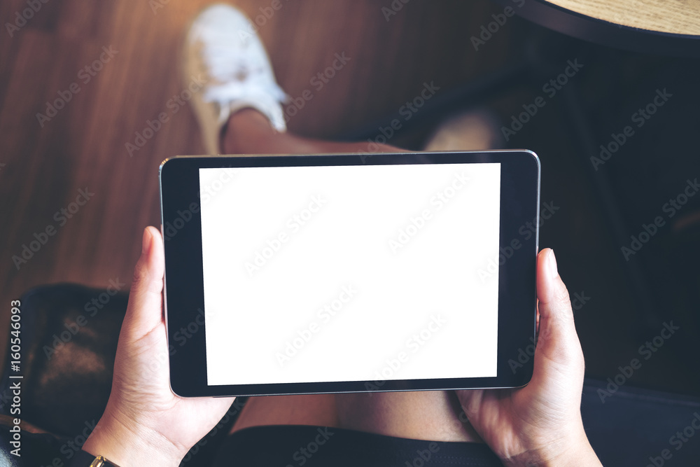Mockup image of woman's hand holding black tablet pc with blank white screen on thigh with wooden floor background in modern cafe