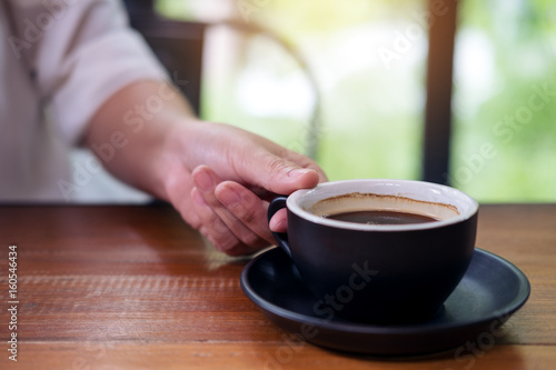 A hand holding a black cup of hot coffee on wooden table in coffee shop