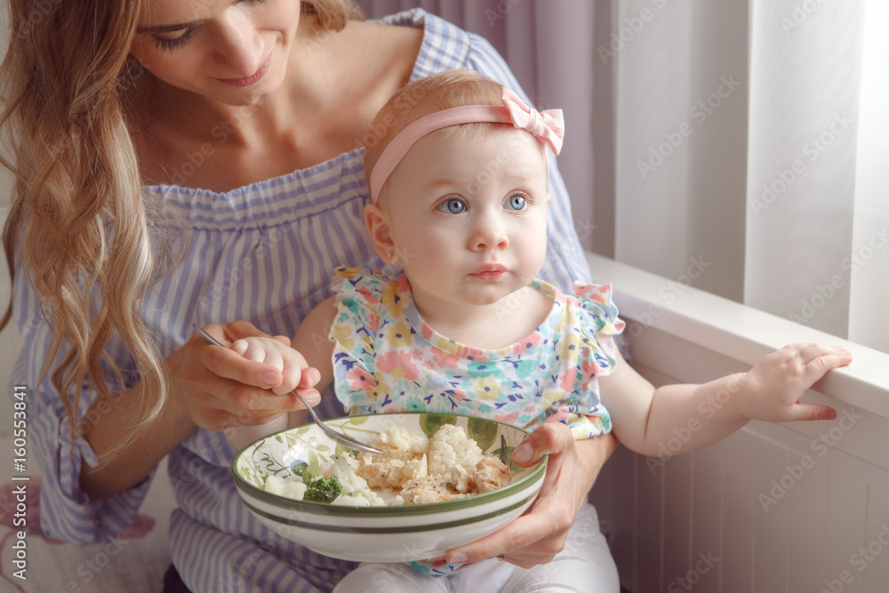 Closeup portrait of young woman mother trying to feed her baby girl daughter with vegetables, broccoli, cauliflower, healthy organic food for child, candid lifestyle family life Closeup portrait of young woman mother trying to feed her baby girl daughter with vegetables, broccoli, cauliflower, healthy organic food for child, candid lifestyle family life