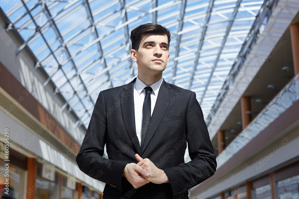 Low angle portrait of successful businessman standing confidently looking away under glass ceiling in modern office building