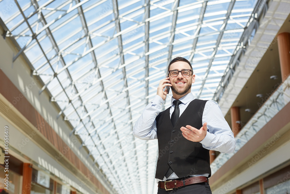Portrait of modern entrepreneur speaking by phone and smiling in office building under glass roof, copy space