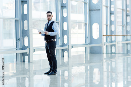 Portrait of contemporary successful businessman holding documents and looking at camera in hall of modern glass hall of office building, copy space