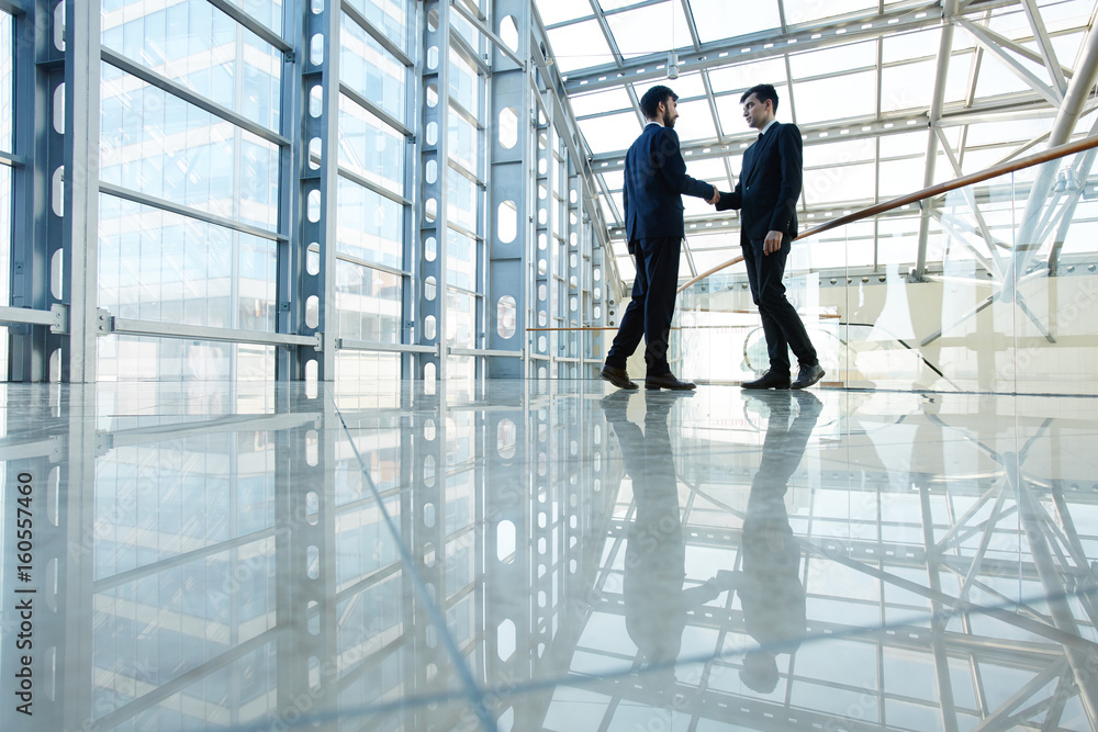 Wide angle portrait of two contemporary business men shaking hands in hall of futuristic glass building Wide angle portrait of two contemporary business men shaking hands in hall of futuristic glass building