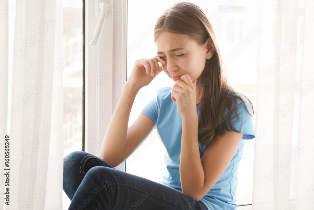 Cute sad girl sitting on window sill at home Cute sad girl sitting on window sill at home