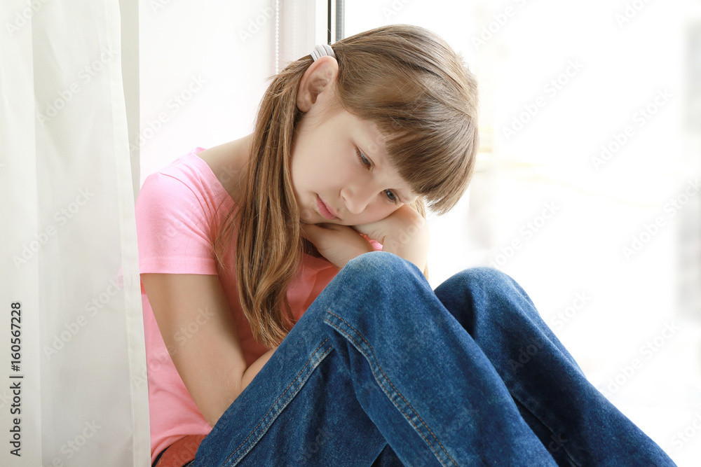 Naklejka premium Sad little girl sitting on window sill at home