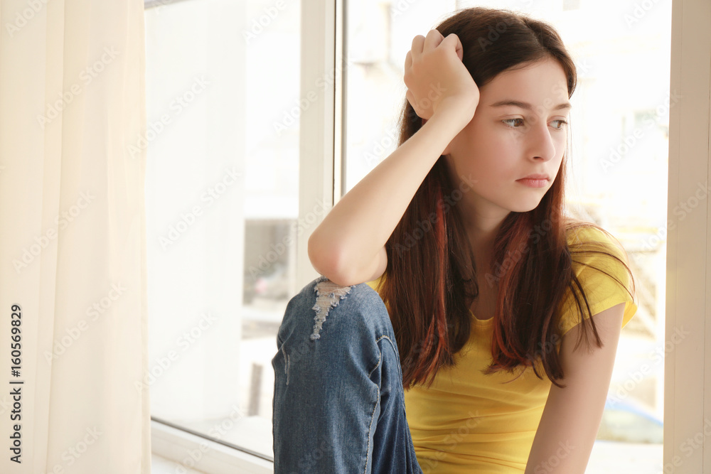Sad pretty girl sitting on window sill at home Sad pretty girl sitting on window sill at home