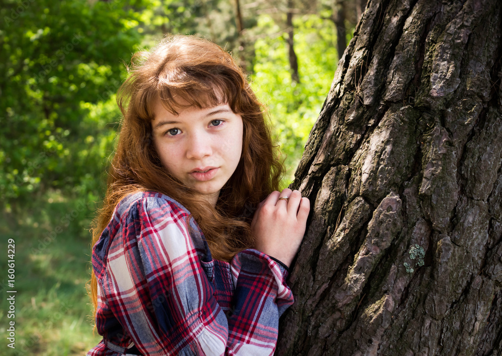 Beautiful red-haired girl sitting under a tree. The wind develops her hair. It is beautifully illuminated by the sun behind. Beautiful red-haired girl sitting under a tree. The wind develops her hair. It is beautifully illuminated by the sun behind.