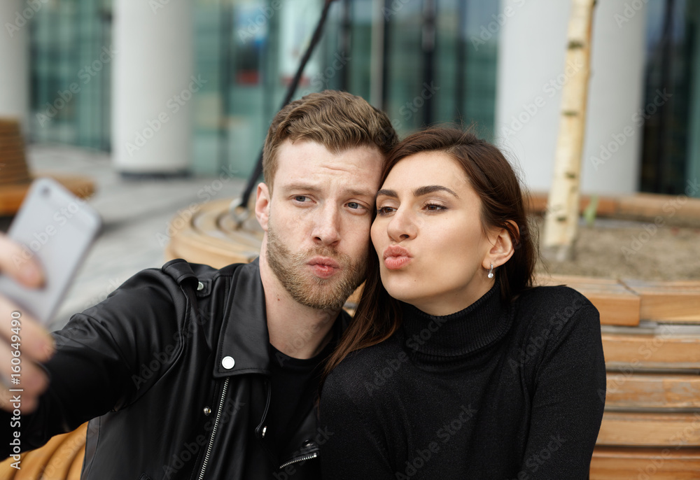 Portrait of attractive fashionable couple taking selfie having fun outdoor. Hipster blonde man in leather jacket with pretty brunette girl sends air kiss looking at screen smart phone. Selective focus
