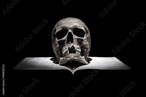 Human skull on old open book on black background under beam of light