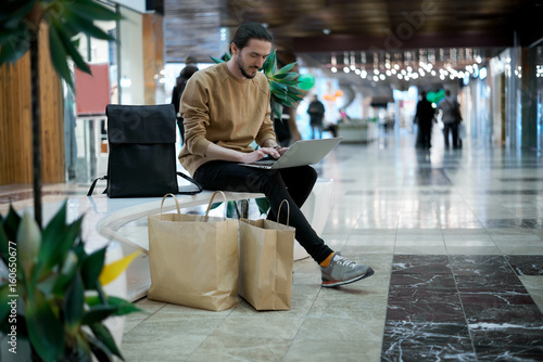 Attractive stylish european student after shopping sitting with notebook and talk friends in social networks. Man buy clothes on summer sales. Guy compare online and real prices.