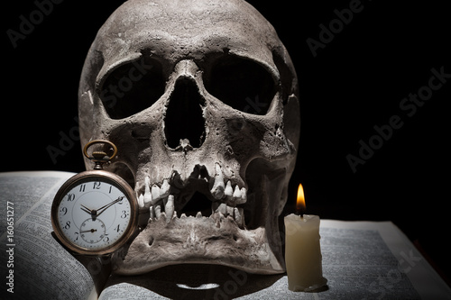 Human skull on old open book with burning candle and vintage clock on black background under beam of light close up
